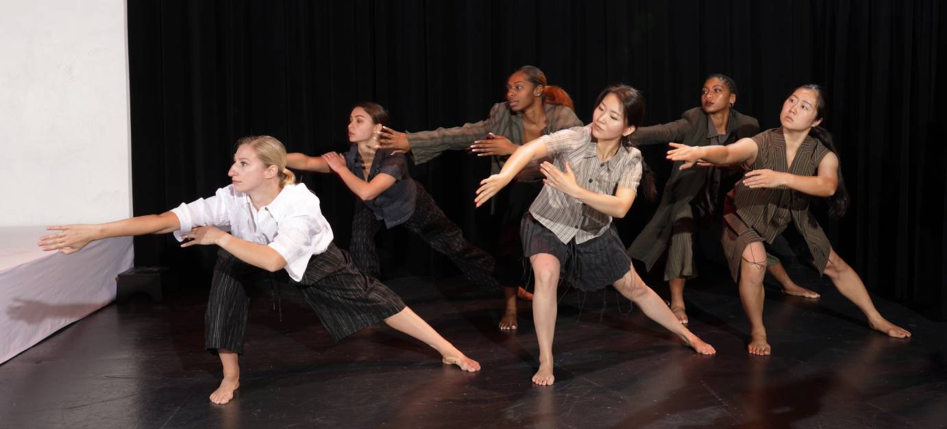 Dancers in shades of gray, white, and beige look over their right shoulder. They extend one arm, turned so that the palm is hidden, and crook the other arm at their waist.